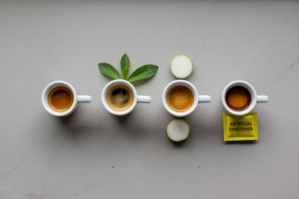 Four identical small white ceramic espresso cups in a row on a light stone surface, each with a different substance beside it: nothing, a small leaf of stevia, a monk fruit, a yellow packet of artificial sweetener.